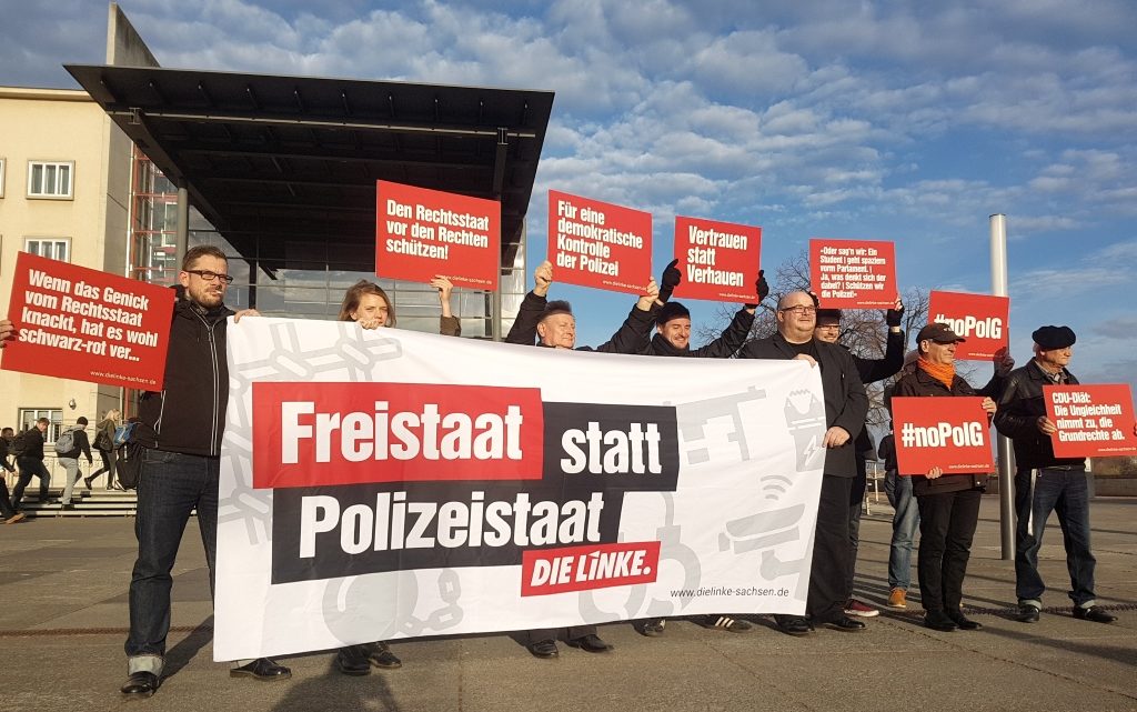 Zu sehen sind mehrere Mitglieder von DIE LINKE. Sachsen, die am 12.11.2018 vor dem Landtag ein Transparent hochhalten, auf dem "Freistaat statt Polizeistaat" steht. Außerdem halten einige noch Protestschilder hoch.