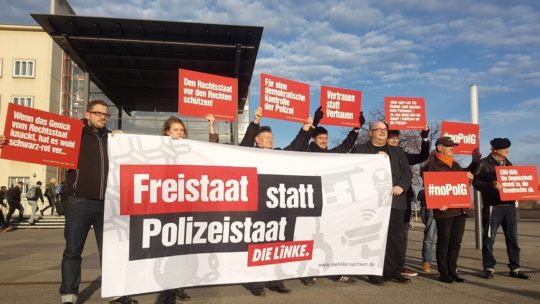Zu sehen sind mehrere Mitglieder von DIE LINKE. Sachsen, die am 12.11.2018 vor dem Landtag ein Transparent hochhalten, auf dem "Freistaat statt Polizeistaat" steht. Außerdem halten einige noch Protestschilder hoch.