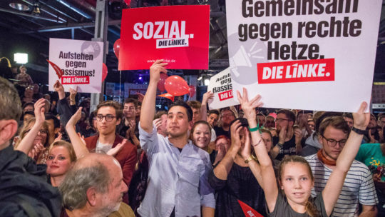 Man sieht Delegierte, die auf einem LINKE-Parteitag Schilder hochhalten. Auf einem steht "Sozial" und auf einem anderen "Gemeinsam gegen rechte Hetze!"