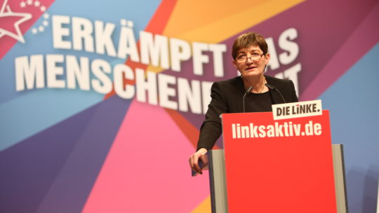 Zu sehen ist Cornelia Ernst bei ihrer Rede auf der VertreterInnenversammlung in Bonn 2019. Hinter ihr steht der geschwungene Schriftzug "Erkämpft das Menschenrecht".