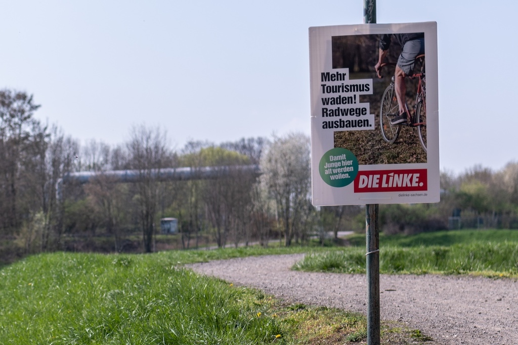 Zu sehen ist ein Wahlplakat mit dem Text: "Mehr Tourismus waden!". Auf dem Plakat sieht man einen Radfahrer, dessen Waden und ein weinrotes Diamantfahrrad. Im Hintergrund ist ein Radweg zu sehen.