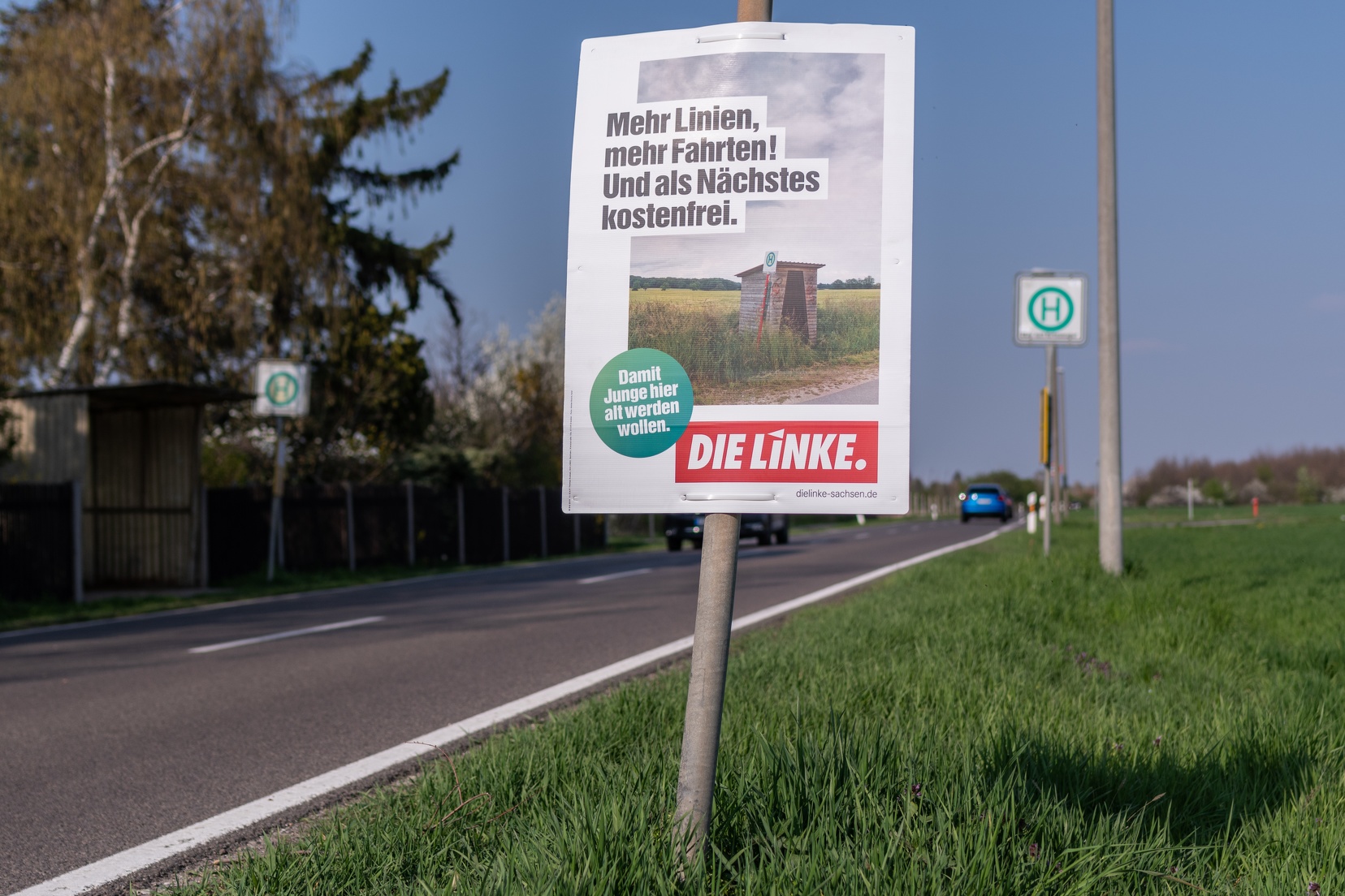 Zu sehen ist ein Wahlplakat mit dem Text: "Mehr Linien, mehr Fahrten! Und als nächstes:Kostenfrei." Das Plakat hängt an einer Landstraße, im Hintergrund ist eine verlassene Bushaltestelle zu sehen.