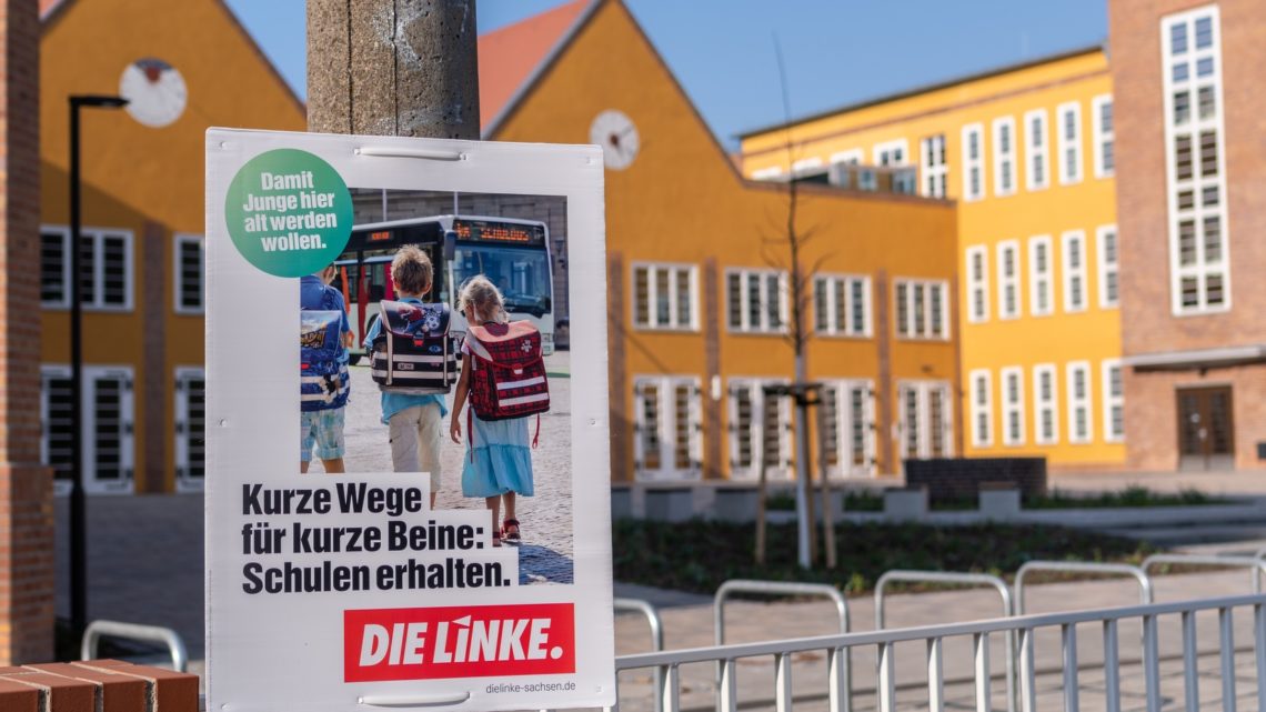 Zu sehen ist ein Wahlplakat mit dem Text: "Kurze Wege für Kurze Beine: Schulen erhalten". Das Plakat hängt vor einer Schule.