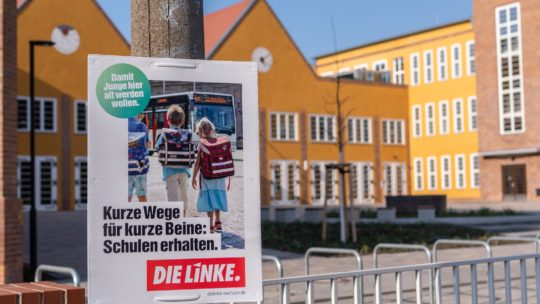 Zu sehen ist ein Wahlplakat mit dem Text: "Kurze Wege für Kurze Beine: Schulen erhalten". Das Plakat hängt vor einer Schule.