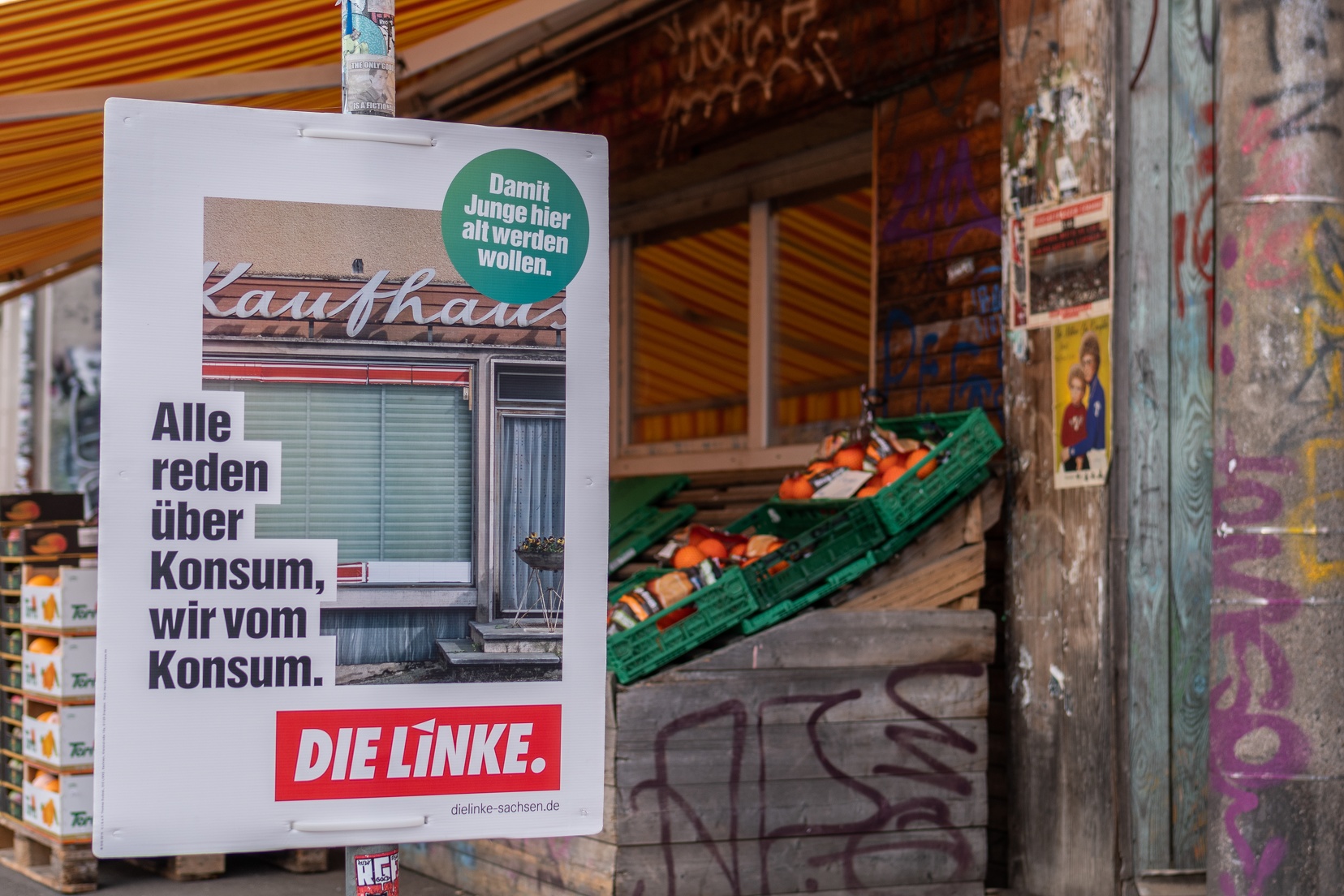 Zu sehen ist ein Wahlplakat mit dem Text: "Alle reden vom Konsum, wir vom Konsum." Zu sehen im Hintergrund ist eine kleine Obstauslage eines Geschäfts.