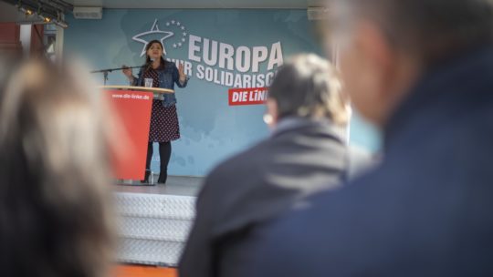 Zu sehen ist ein Bild vom Europawahlkampf 2019. Im Hintergrund sieht man die Rücken von Zuhörenden, im Hintergrund spricht Özlem Demirel auf einer Bühne.
