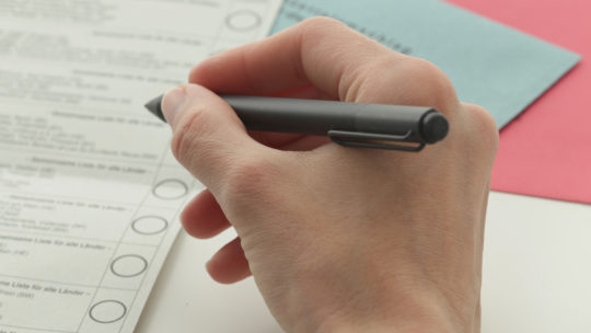Zu sehen ist, wie jemand einen Stift hält und darunter Wahlzettel liegen.