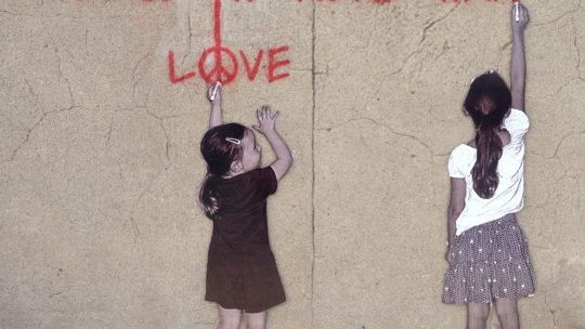 Graffiti bei dem zwei Mädchen an eine Wand "Please no more war, love" schreiben