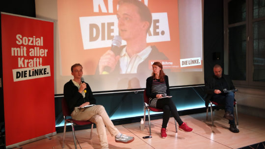 Foto von der Wasserstoffkonferenz. Zu sehen ist das Podium mit Marco Böhme und Antonia Mertsching. Links steht ein Roll-Up mit dem Text "Sozial mit aller Kraft. DIE LINKE"