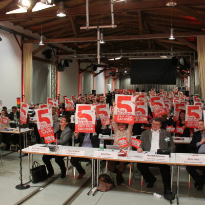 Auf einem Landesparteitag von DIE LINKE. Sachsen halten die Delegierten Plakate hoch, auf denen "5 Tage Bildungszeit für Sachsen" steht.