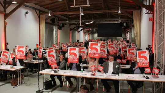 Auf einem Landesparteitag von DIE LINKE. Sachsen halten die Delegierten Plakate hoch, auf denen "5 Tage Bildungszeit für Sachsen" steht.