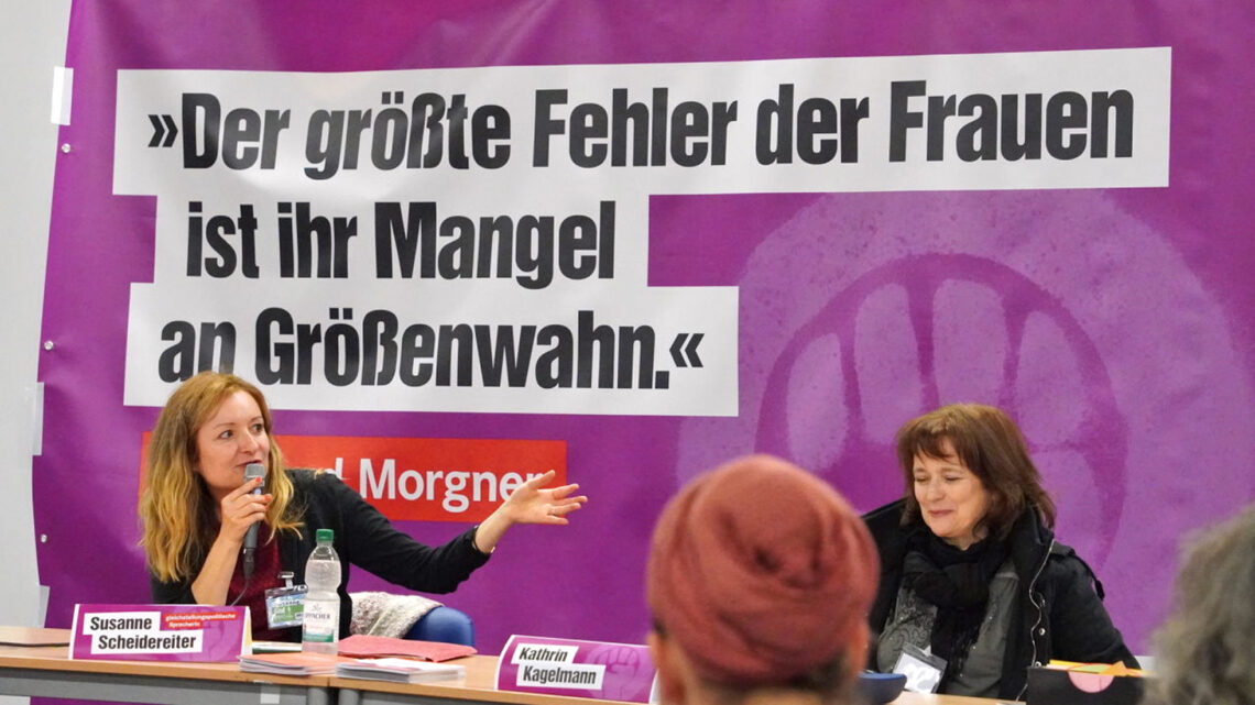 Foto von der Tagungsleitung des Finta*plenums. Im Hintergrund ist ein Banner mit dem Text: "Der größte Fehler der Frauen ist ihr Mangel an Größenwahn"