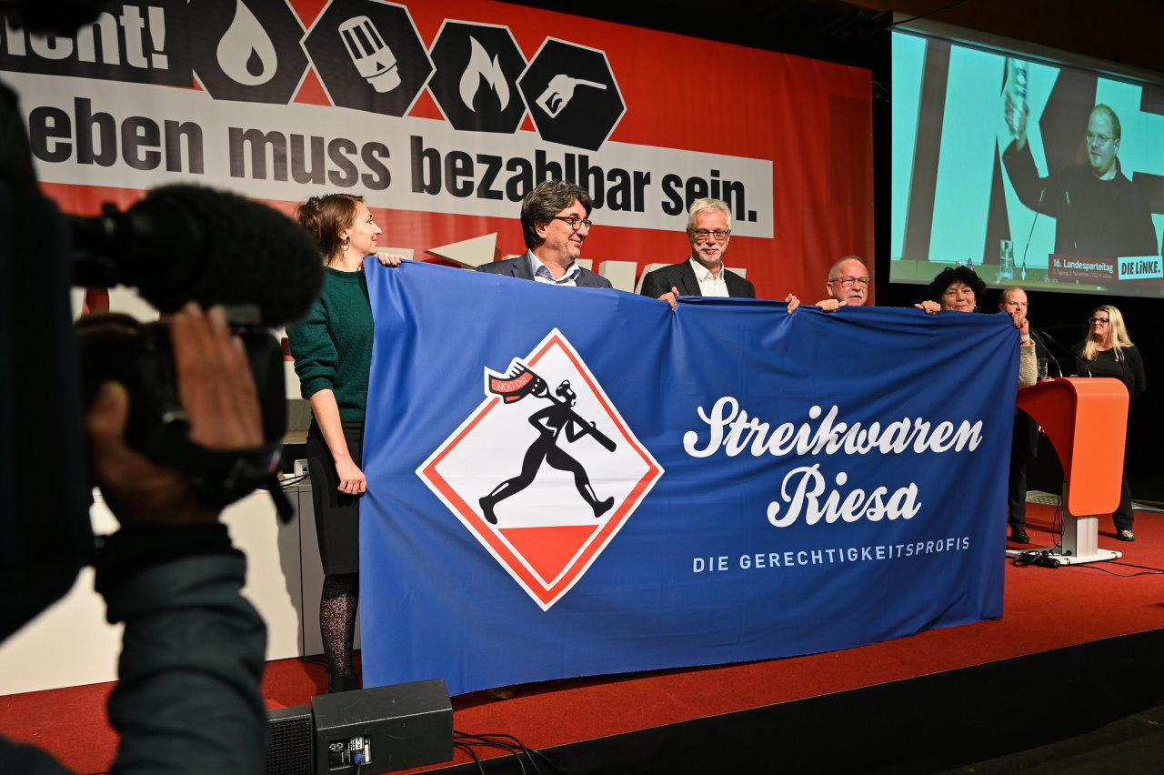 Zu sehen sind Anna Gorskih, Stefan Hartmann, Markus Schlimbach und zwei Genoss*innen aus Meißen die ein Banner "Streikwaren Riesa - die Gerechtigkeitsprofis" auf einer Bühne des Landesparteitages halten.