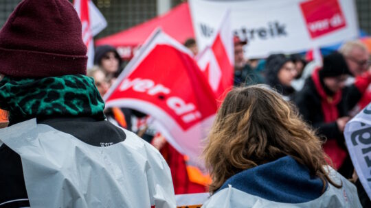 Foto von einem Streik. Man sieht eine ver.di-Fahne und eine Frau von hinten, die eine ver.di-Weste trägt.