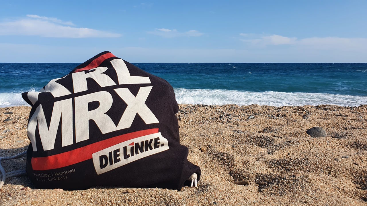 Schwarzer Turnbeutel mit dem Aufdruck "KRL MRX" und Logo DIE LINKE an einem Sandstrand am Meer