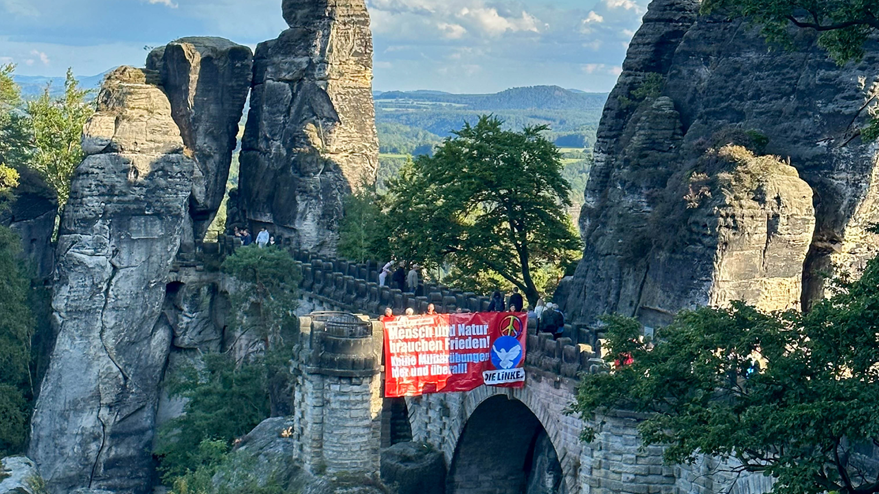Ein Foto der Basteibrücke in der sächsischen Schweiz, von der ein großes rotes Banner mit dem Text „Mensch und Natur brauchen Frieden!“ runter hängt.